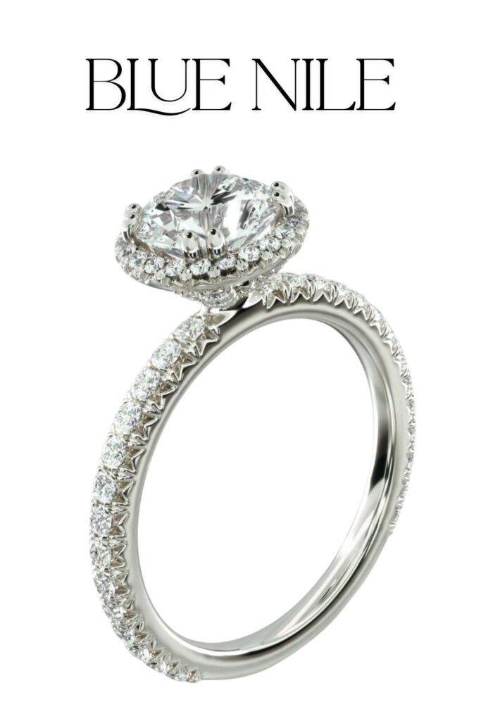 The Blue Nile Studio Heiress Halo Diamond Engagement Ring features a stunning round diamond, elegantly set in a halo of smaller diamonds, showcasing its brilliant cut and high clarity. The band, crafted from polished platinum, is adorned with a row of pavé-set diamonds, enhancing its luxurious appeal. This exquisite design exemplifies timeless elegance and meticulous craftsmanship.