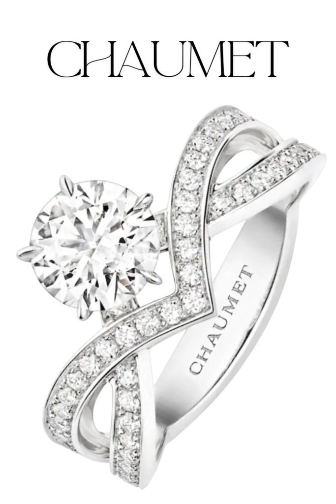 The Chaumet Joséphine Amour d'Aigrette solitaire engagement ring features a stunning round brilliant diamond, set in a polished platinum band embellished with pavé-set diamonds along its twisting shoulders. The design showcases exquisite craftsmanship, harmonizing contemporary elegance with classic bridal allure. This luxurious piece epitomizes the beauty and sophistication associated with the Chaumet brand.
