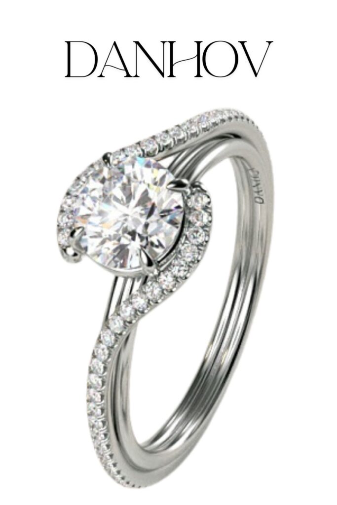 The Danhov Abbraccio Handmade Diamond Engagement Ring features a stunning, round brilliant diamond with excellent cut, clarity, and color, securely set in a graceful solitaire mounting. The band is crafted from 14k white gold, elegantly entwined, adorned with pavé-set diamonds that enhance its brilliance and sophistication. This ring beautifully exemplifies the exquisite craftsmanship and modern design aesthetic synonymous with the Danhov brand.