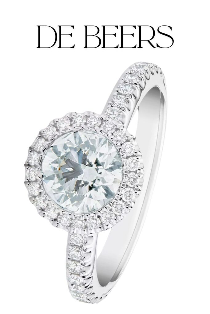 The De Beers Aura ring features a stunning round brilliant diamond at its center, enhanced by a halo of smaller diamonds that showcases exceptional clarity and color. Set in lustrous platinum, the band is adorned with pavé-set diamonds along its shoulders, adding sophistication to the solitaire design. This elegant piece exemplifies high-end craftsmanship and timeless beauty.