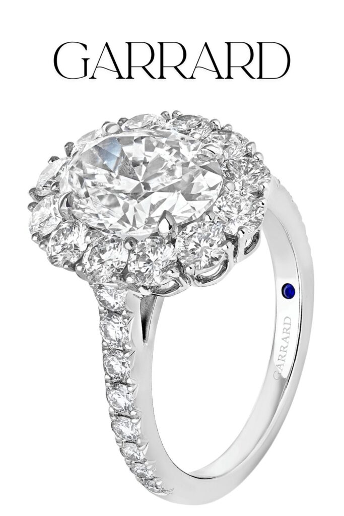 The Garrard Jewelled Vault engagement ring features a stunning 3.01-carat oval diamond at its center, displaying exceptional clarity and color. Set in a luxurious platinum band, the diamond is surrounded by a halo of smaller diamonds, enhancing its brilliance with a pavé setting along the band. A small blue stone is subtly embedded inside, adding a unique touch to this exquisite piece.