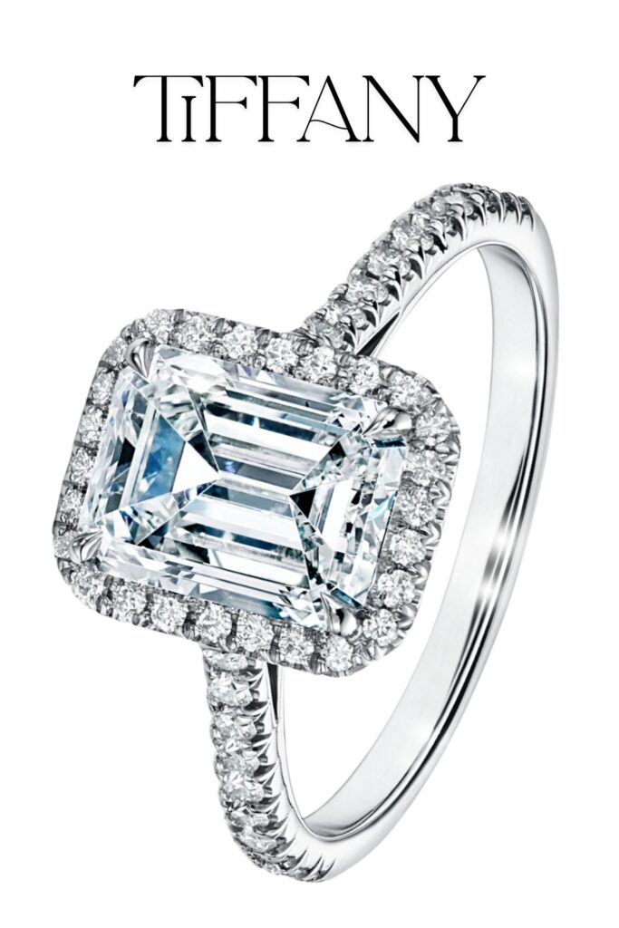 The Tiffany Soleste Emerald-cut Halo Engagement Ring features a stunning emerald-cut diamond at its center, surrounded by a halo of sparkling round diamonds, all meticulously set in a platinum band. The ring showcases exceptional clarity and color, enhancing its luxurious design and craftsmanship. The band is adorned with delicate pavé diamonds, adding to the overall brilliance and elegance of this exquisite piece.