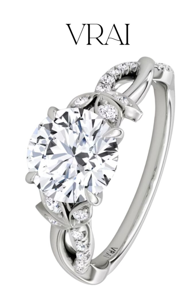 The Vrai Twisted Floral Engagement Ring features a brilliant round diamond at its center, showcasing exceptional clarity and color, elegantly set within a twisted floral design. The band is crafted from lustrous white gold and adorned with delicate pavé diamonds along the shoulders, enhancing its refined sophistication.