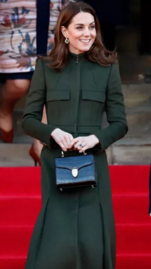 Kate Middleton is wearing the Aspinal of London Midi Mayfair handbag in Evergreen Patent Croc, which complements her tailored, olive green coat featuring a high neckline and structured silhouette. The bag's sleek and compact design, accented with a gold clasp, adds a sophisticated touch to her ensemble, while its deep color harmonizes with the coat, creating a cohesive and polished appearance. The overall look is elegantly finished with understated jewelry and a classic hairstyle, emphasizing her refined style.