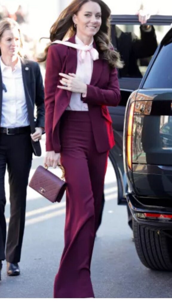 The handbag is a Chanel Classic Flap Bag in burgundy quilted leather, featuring an elegant enamel handle that adds a touch of sophistication to Kate Middleton's look. Complementing her tailored burgundy suit, which consists of a fitted blazer and high-waisted trousers, the bag enhances her poised silhouette while maintaining a cohesive color scheme. The overall style exudes modern elegance, accentuated by the contrasting soft pink bow tie at the neck.