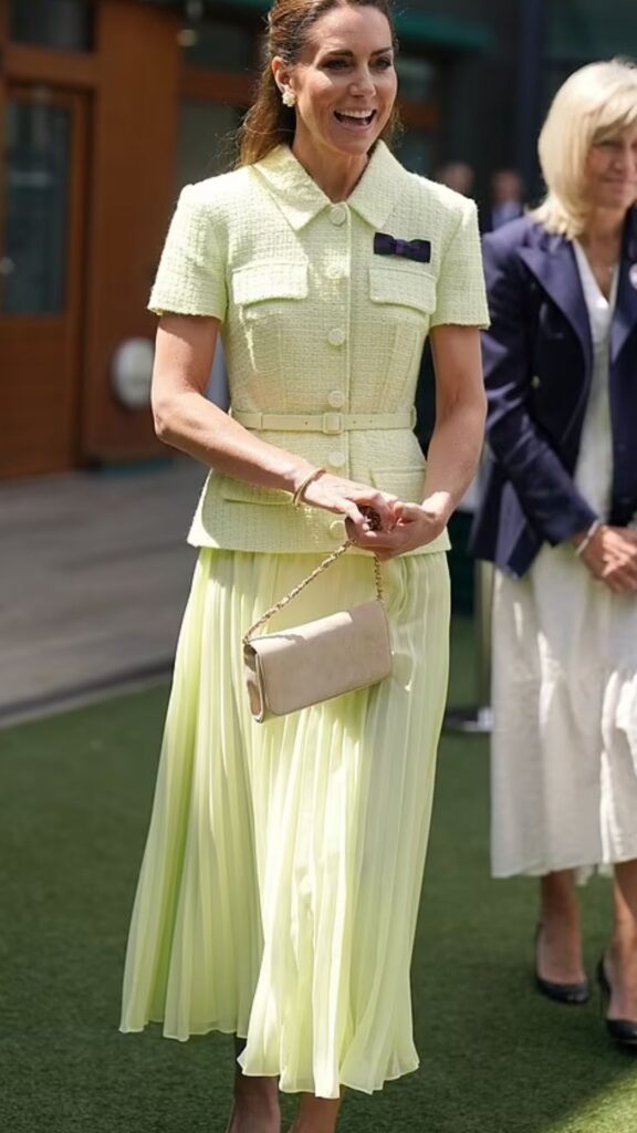 Kate Middleton pairs the Emmy London Natasha Clutch in blush suede with a light yellow, textured jacket and pleated skirt ensemble. The elegant handbag mirrors the soft color palette and enhances the sophistication of her structured top, while its sleek silhouette and chain detailing add a modern touch to the overall look. This combination reinforces her signature style of refined femininity with a contemporary edge.