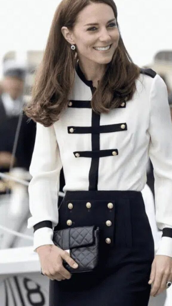 The Jaeger Kate Bag in navy quilted leather complements Kate Middleton's sophisticated look, featuring a stylish white and black ensemble with gold buttons. The bag, with its structured silhouette, adds a touch of elegance while balancing the boldness of the fitted attire. Its rich color harmonizes with her tailored outfit, enhancing the overall chic appearance.