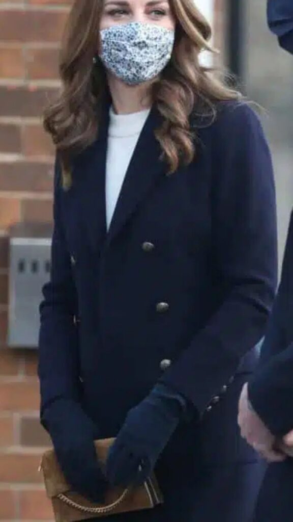 The image features Kate Middleton wearing a tailored navy double-breasted coat over a light turtleneck, complemented by navy gloves. The Metier Roma small brown suede shoulder bag adds warmth and elegance to her ensemble, contrasting beautifully with the navy hues while its subtle chain detail enhances the sophisticated feel of her overall look.