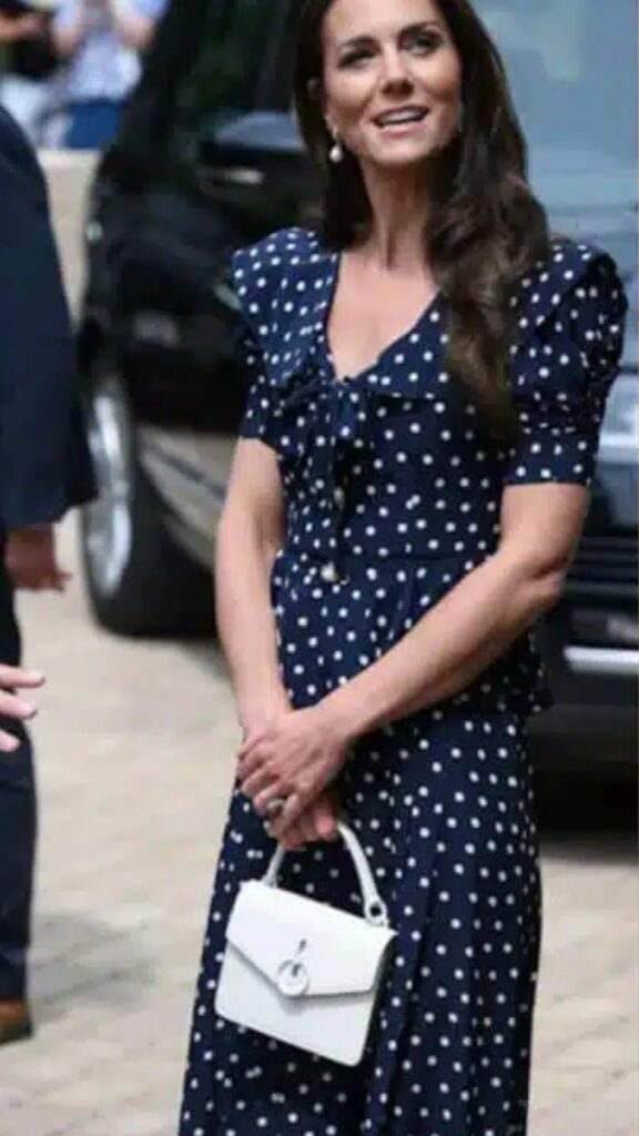 Kate Middleton's outfit features a fitted navy dress adorned with white polka dots, a flattering V-neckline, and short puff sleeves that emphasize her silhouette. She completes the look with a Mulberry Amberley Small Crossbody Bag in white, which adds a contemporary touch and contrast to her ensemble, enhancing the overall elegance of her appearance. The handbag's structured shape and clean lines complement the playful design of the dress while providing a chic accessory for her outing.