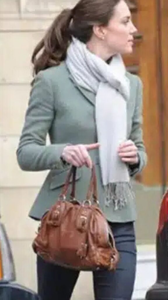 Kate Middleton is seen carrying a Prada tan bowler bag, which adds a polished contrast to her casual-chic ensemble. The structured silhouette of the bag complements her tailored green blazer and slim-fit trousers, while its rich brown hue harmonizes with the soft gray scarf, enhancing the overall sophistication of her look. The handbag’s versatility makes it an ideal accessory for her effortlessly elegant style.