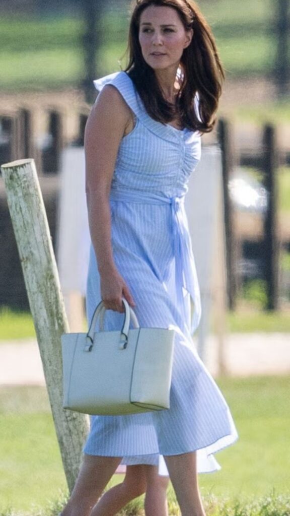 The Victoria Beckham Quincy Bag in white adds an elegant touch to Kate Middleton's light blue, striped dress, which features a fitted bodice and flowing skirt that gracefully drapes past the knees. The bag's structured silhouette complements the dress's playful ruffled sleeves and soft color palette, enhancing the overall sophisticated yet relaxed vibe of the look. The accessories are thoughtfully paired, further emphasizing her refined style.