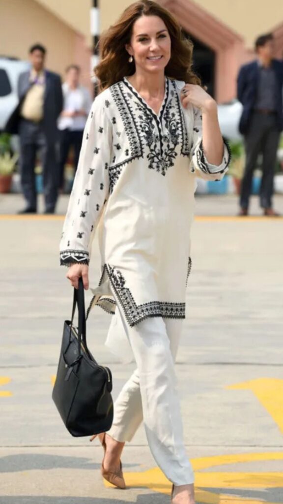 Kate Middleton complements her elegant white tunic adorned with intricate black embroidery by carrying a Smythson Panama East West Zip Tote, which adds a modern touch to her traditional look. The sleek black leather contrasts beautifully with her outfit and provides a sophisticated yet functional accessory, fitting seamlessly with her tailored white trousers and nude heels. This combination emphasizes her stylish approach while ensuring ease of movement and practicality.