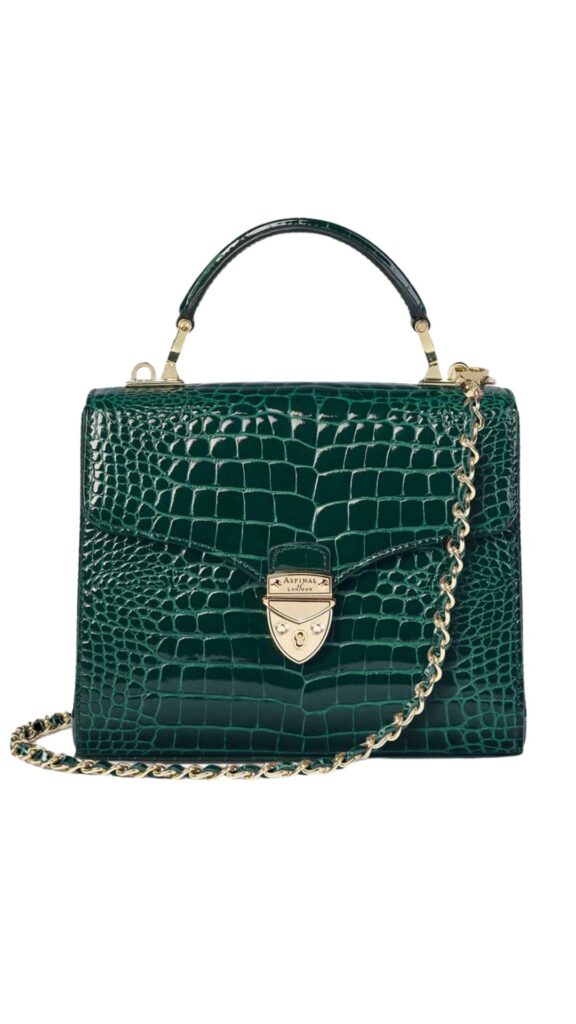 The Aspinal of London Midi Mayfair bag features a structured silhouette with a polished evergreen patent finish that mimics crocodile texture. It is adorned with a sleek gold clasp and a detachable gold chain strap, adding versatility for both handheld and shoulder wear. This elegant handbag combines sophisticated style with practical functionality.