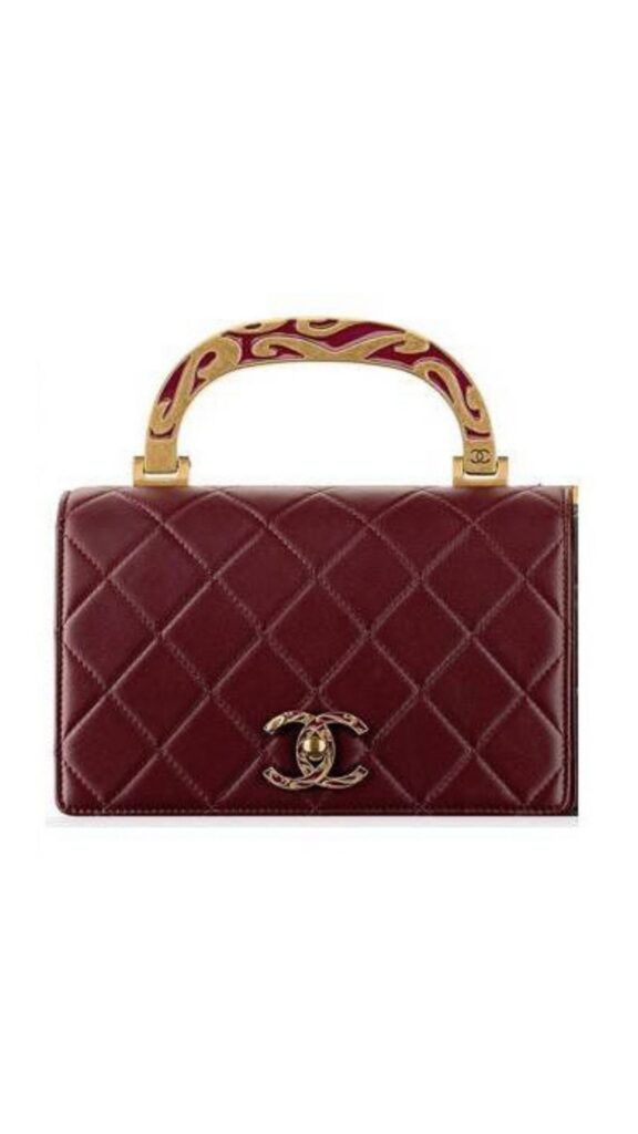 The Chanel Classic Flap Bag features a rectangular silhouette crafted from burgundy quilted leather, accented by a gold-tone enamel handle with a decorative design. A signature interlocking CC logo sits prominently on the front, adding a luxurious touch. This stylish piece combines elegance and functionality, making it a versatile accessory for various occasions.