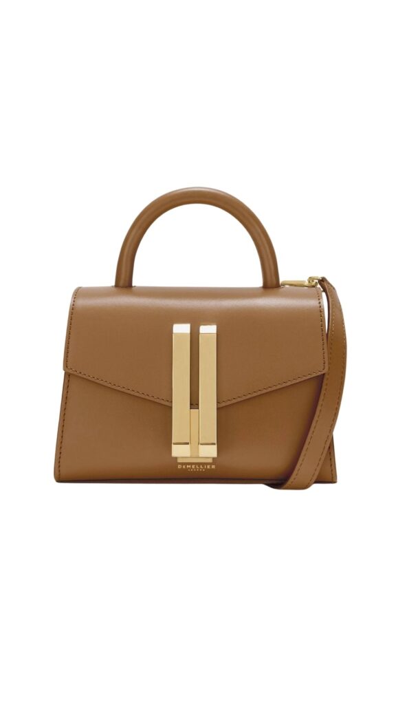 The Demellier Nano Montreal handbag features a structured silhouette with a top handle and a detachable shoulder strap for versatile carrying options. It is crafted from smooth deep toffee leather and accented with a striking gold-tone clasp closure at the front. The clean lines and elegant design make it a chic accessory suitable for various occasions.