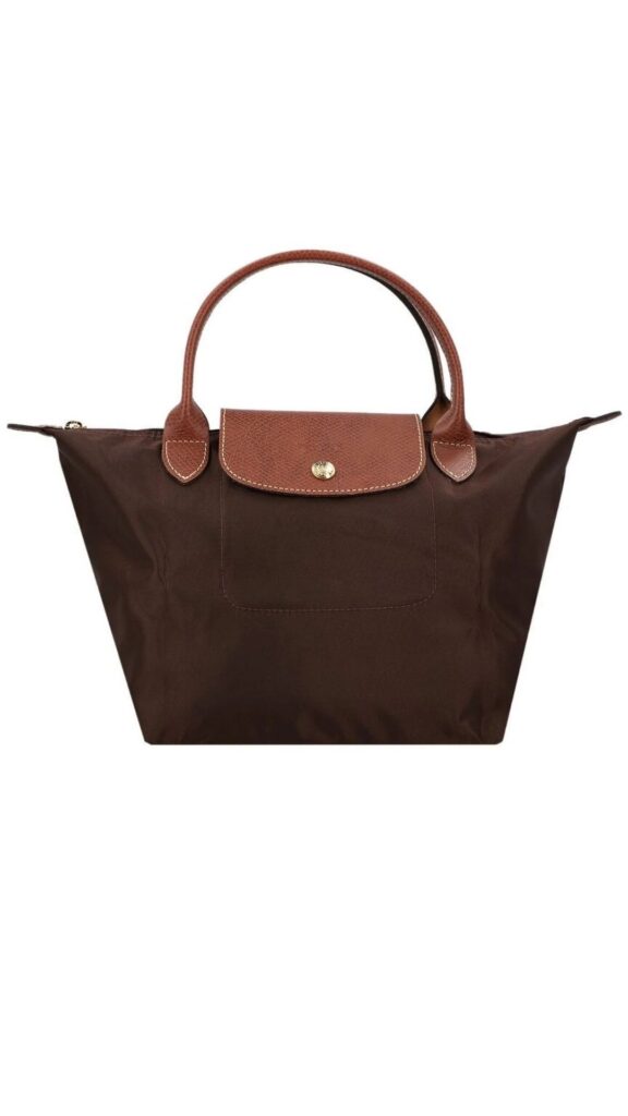 This Longchamp Le Pliage Small handbag features a trapezoidal silhouette with a spacious canvas body and sturdy leather handles. It is primarily brown with a smooth leather flap and accents in a darker shade of brown. The design includes a gold snap closure at the top and a front pocket for added functionality.