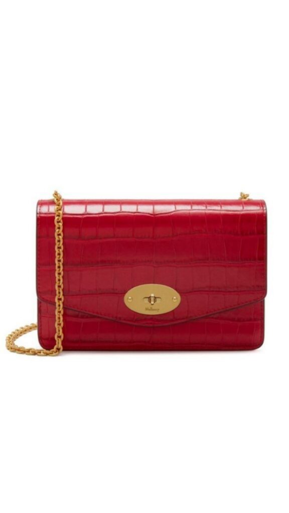 The Mulberry Small Darley Bag in red croc features a sleek rectangular silhouette crafted from textured red leather with a crocodile print. It includes a gold-tone chain strap for versatile wearing options and is adorned with a signature gold-colored clasp at the front. This chic handbag combines elegance and modern appeal, perfect for various occasions.