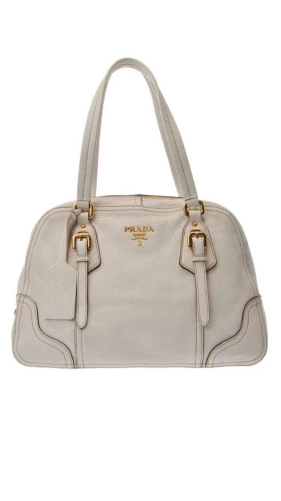 The Prada White Boston Bag features a structured silhouette with dual top handles and elegant gold-tone hardware. Crafted from soft, pebble-grain leather, it is presented in a soft white hue with subtle stitching details. The bag includes adjustable side straps, enhancing its stylish and versatile design.