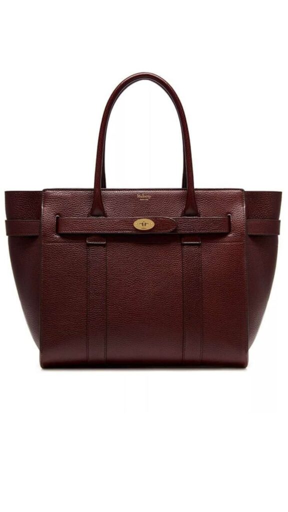 The Mulberry Bayswater Zipped Tote features a structured silhouette with dual top handles, crafted from rich burgundy leather. It showcases a distinctive, elegant design accentuated by a central leather strap and a gold-tone logo plaque, providing both style and functionality. This versatile tote is ideal for everyday use, offering ample space for personal items.