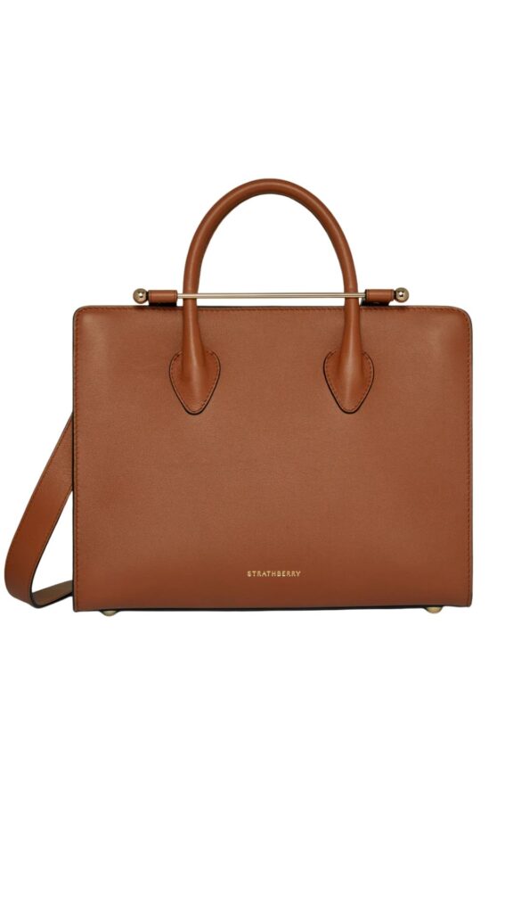 The Stratberry Midi Tote in Chestnut features a structured silhouette with a sleek design, showcasing a warm chestnut brown leather exterior. It is accented by a gold-tone metal handle and distinctive pointed details on the sides, providing both elegance and functionality. This versatile handbag can be carried by hand or worn crossbody with its adjustable strap.
